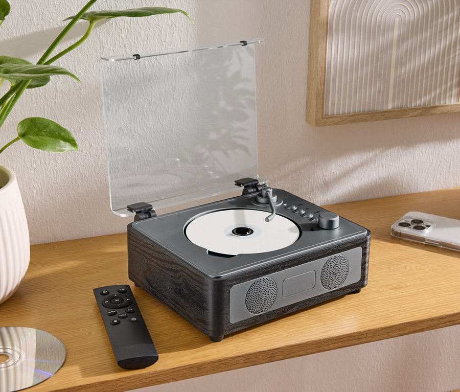 Auf einem Tisch steht ein CD/Bluetooth®-Lautsprecher mit einer CD, einer Fernbedienung und einem Handy.