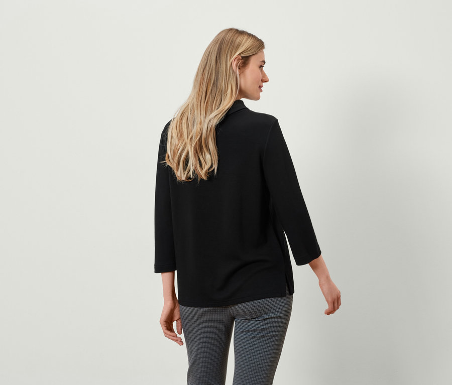 Blonde Frau von hinten in schwarzem Oberteil mit Dreiviertelärmeln und grauer karierter Hose.