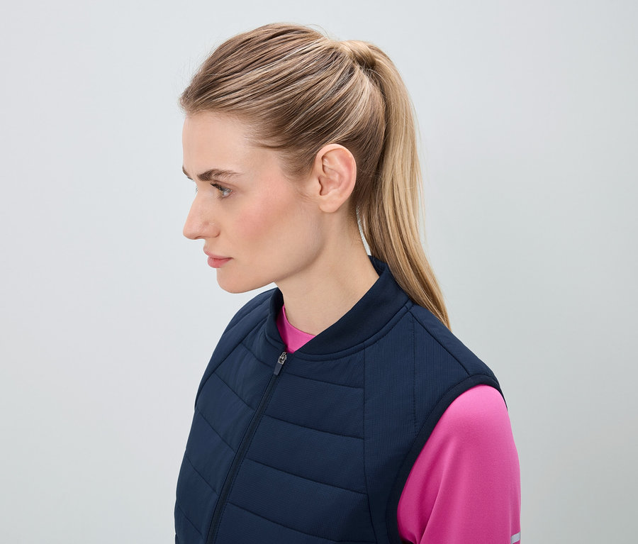 Profil einer Frau mit blonden Haaren, die zu einem Pferdeschwanz zusammengebunden sind, gekleidet in einer dunkelblauen Steppweste und einem rosa Top.