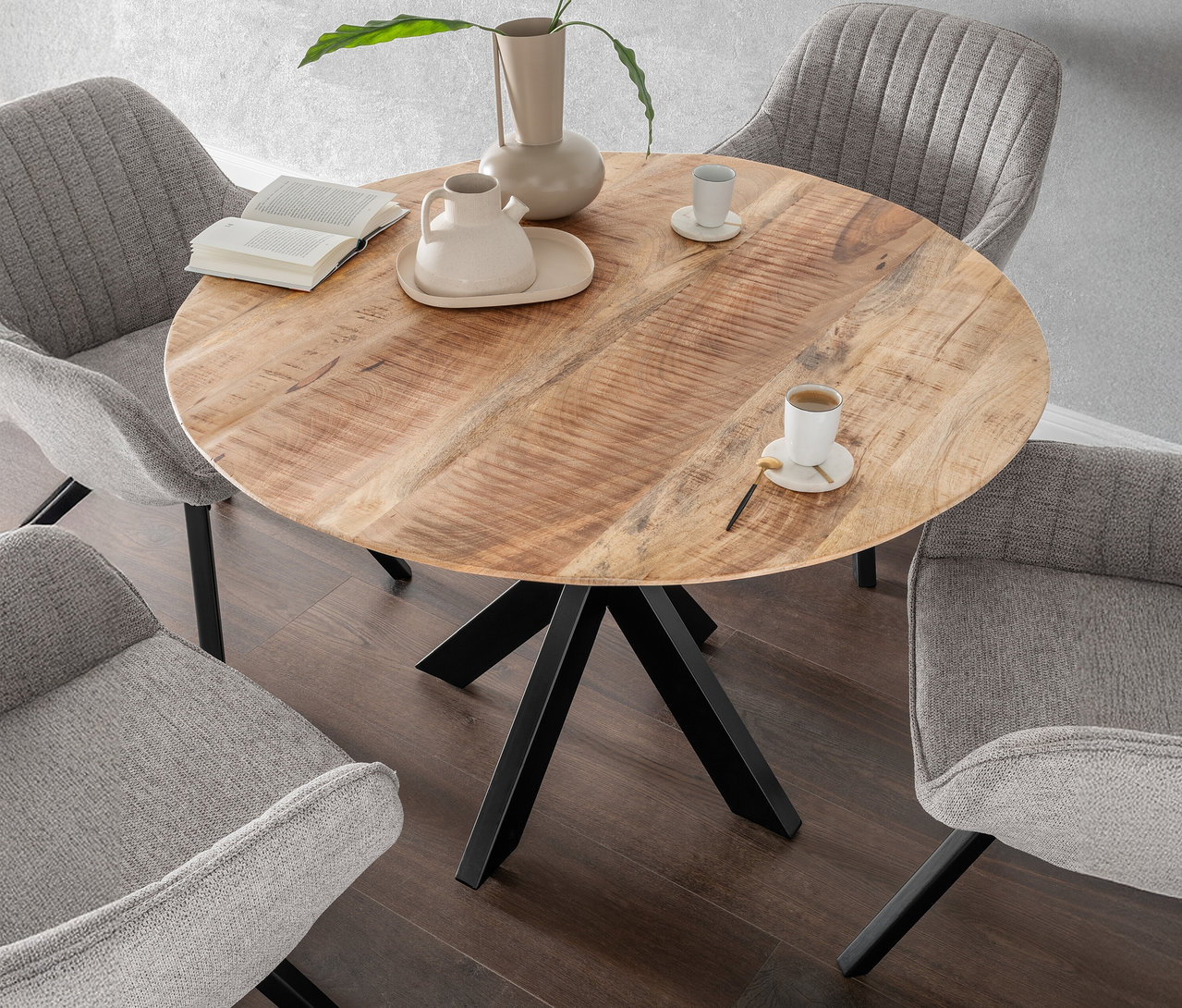 Ein runder Holztisch mit vier grauen Stühlen, einer Vase, einer Karaffe, Tassen und einem Buch.