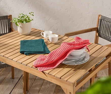 Auf einem Holztisch stehen zwei weiße Tassen, ein grünes und ein rosa Waffeltuch sowie Teller und eine Schüssel.