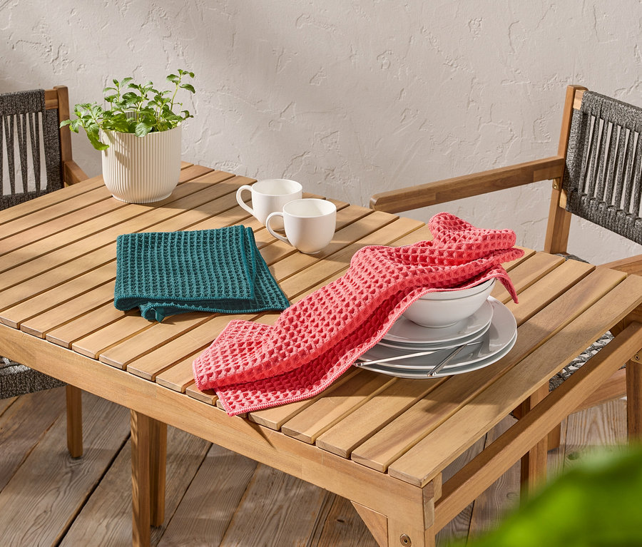 Auf einem Holztisch stehen zwei weiße Tassen, ein grünes und ein rosa Waffeltuch sowie Teller und eine Schüssel.