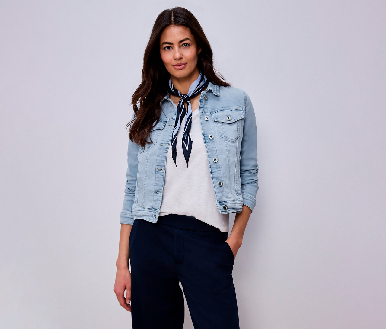 Frau mit hellblauer Jeansjacke, weißem T-Shirt, marineblauer Hose und blau-weiß gestreiftem Halstuch vor hellem Hintergrund.