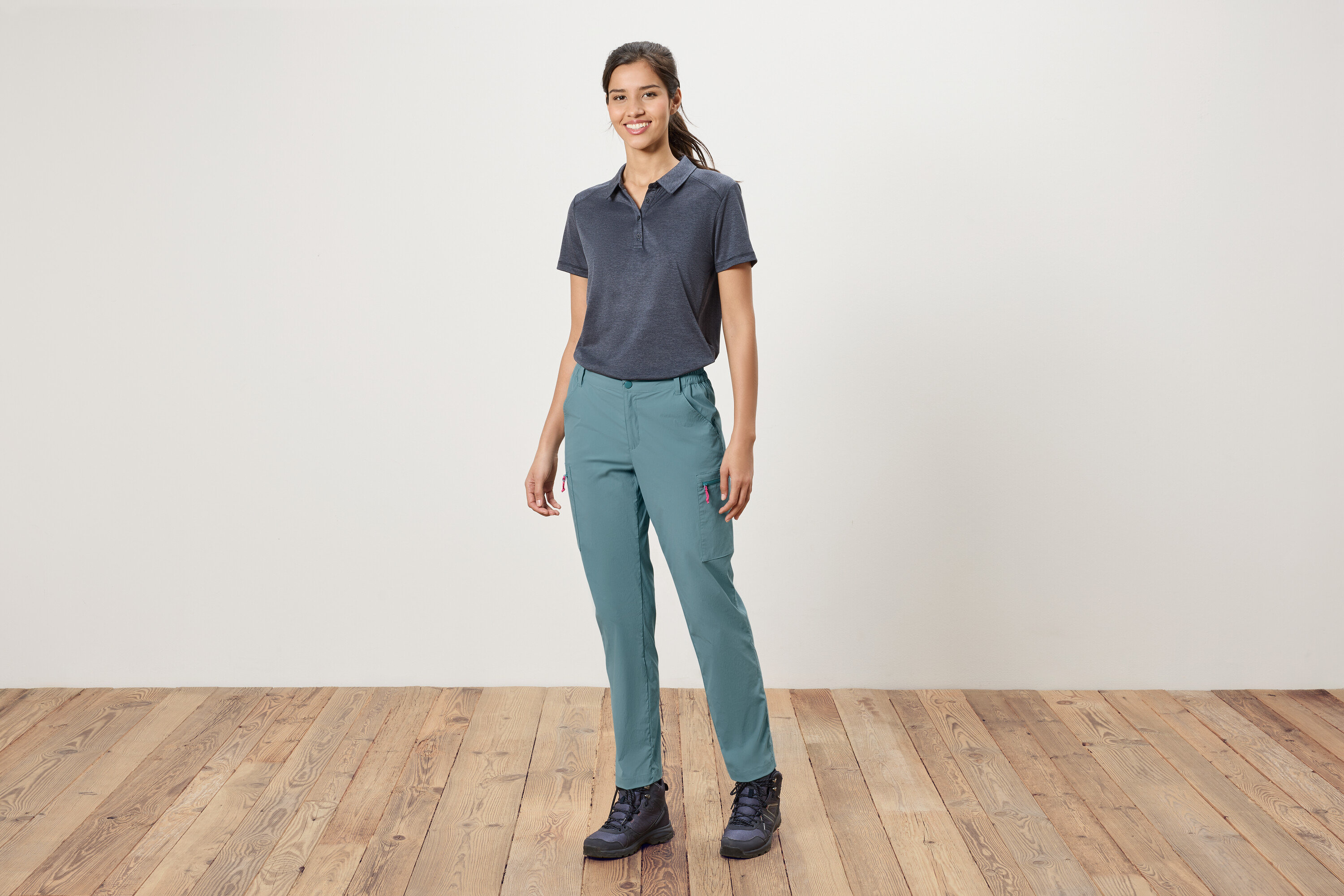 Eine Frau steht vor einer weißen Wand und trägt ein blaues Polohemd, blaue Hosen und Wanderschuhe.