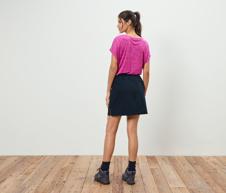 Eine Frau mit Pferdeschwanz steht mit dem Rücken zum Betrachter und trägt ein pinkes T-Shirt, einen schwarzen Rock und graue Schuhe.