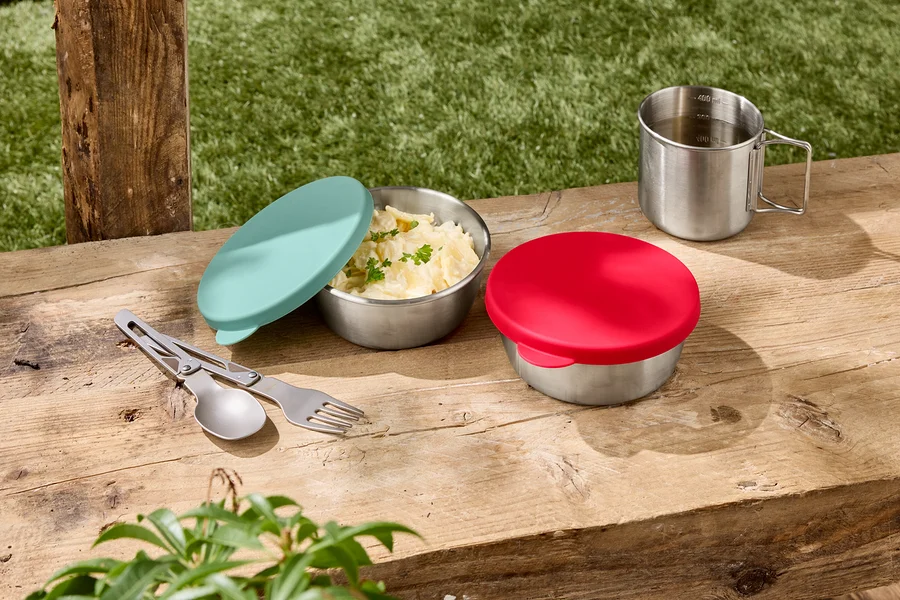 Zwei Edelstahlschüsseln mit Deckel, eine davon mit Kartoffelsalat, daneben ein silberner Becher und ein Besteckset auf einem Holztisch.