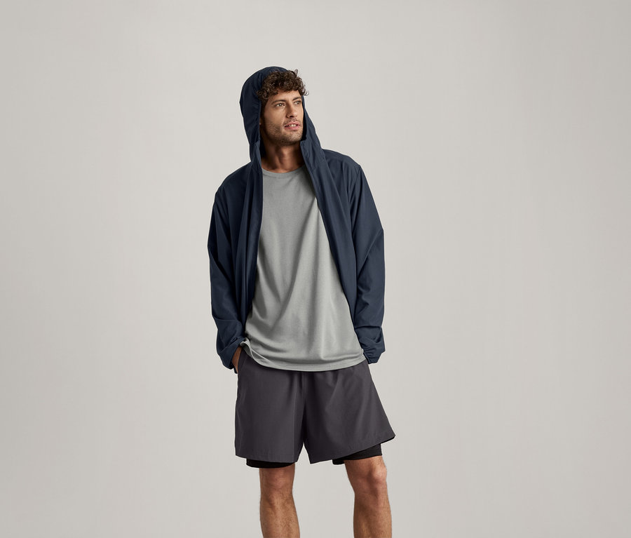 Mann trägt ein hellgrau meliertes Sportshirt, eine dunkelblaue Jacke mit Kapuze und Shorts.