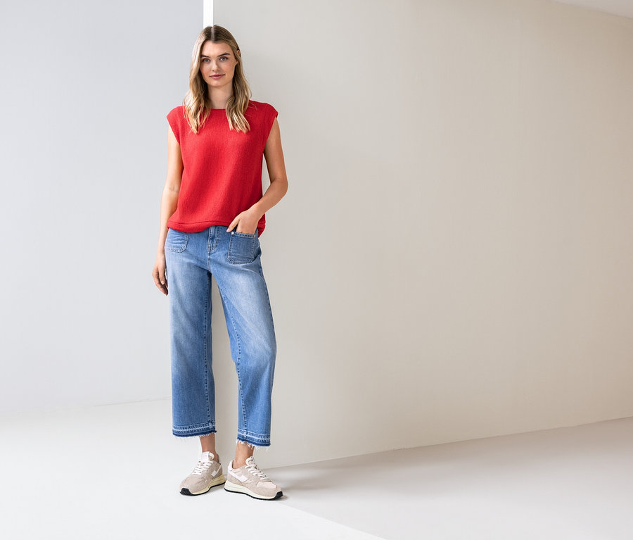 Frau steht in einem hellen Raum, trägt ein rotes Top, blaue Jeans und Turnschuhe.