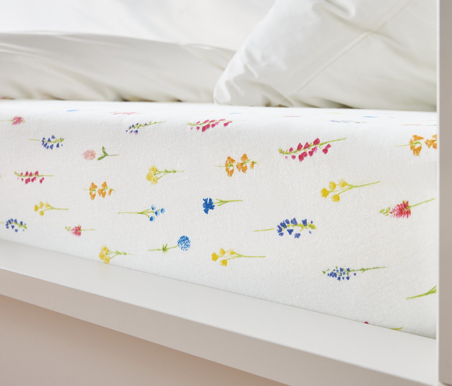 Nahaufnahme eines Bettes mit weißem Bettlaken, das mit einem Blumenmuster in Gelb, Blau, Orange und Rosa bedeckt ist.