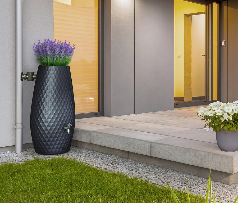 Eine schwarze Regentonne mit Rautenmuster und lila Blumen steht neben einem Fallrohr und einer Haustür.