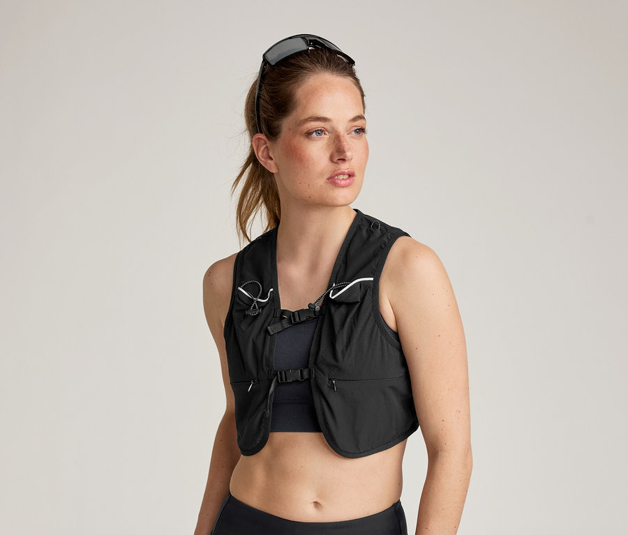 Frau mit Sonnenbrille auf dem Kopf trägt eine schwarze Laufweste über einem Sport-BH und Leggings.