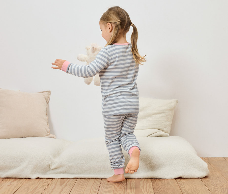 Mädchen in grau-weiß gestreiftem Pyjama hält Teddybär auf einer Couch.
