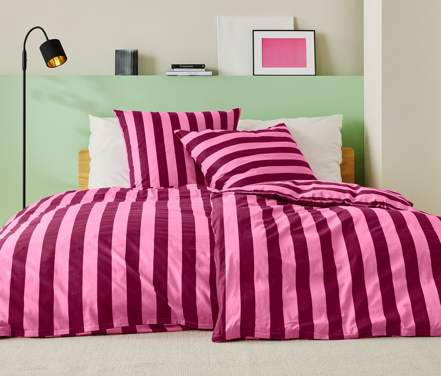 Bett mit rosa-weinroter Streifenbettwäsche, zwei Kissen und einer Stehlampe.