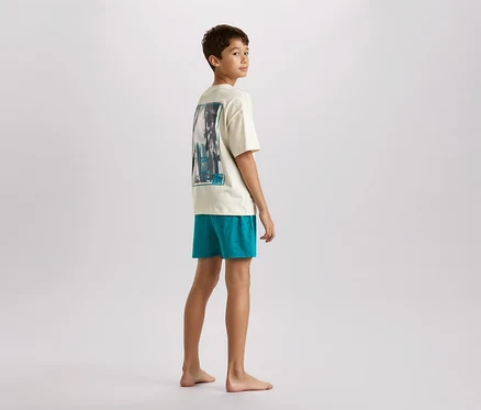 Ein junger Teenager steht barfuß und blickt über die Schulter. Er trägt ein cremefarbenes T-Shirt mit einem Aufdruck und türkisfarbene Shorts.