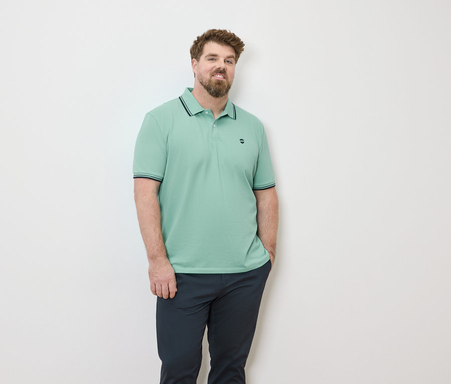 Mann mit Bart, der ein hellgrünes Poloshirt und eine dunkle Hose vor einer weißen Wand trägt.