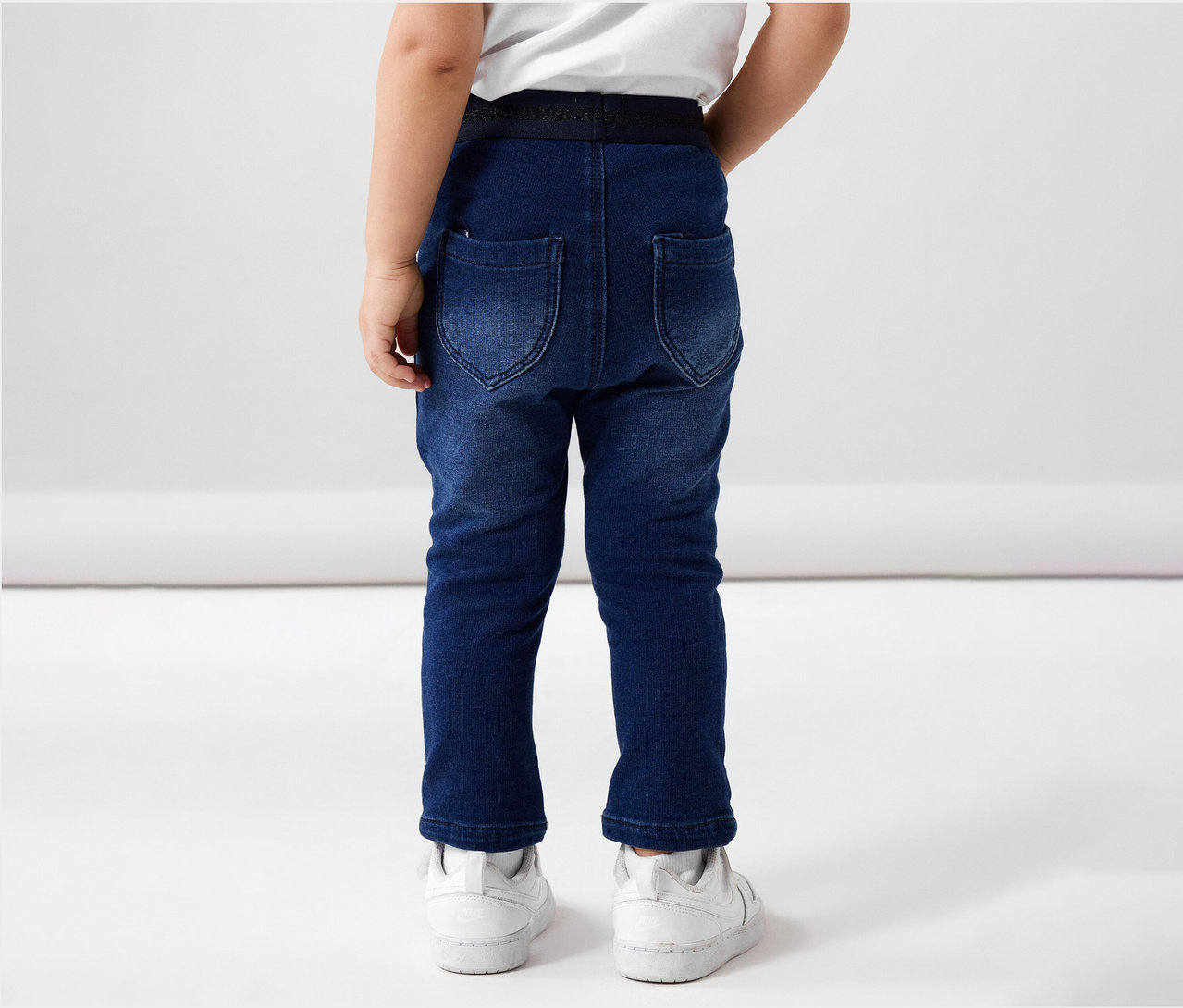 Rückansicht eines Kindes in NAME IT Kinder-Sweat-Jeans und weißen Turnschuhen.