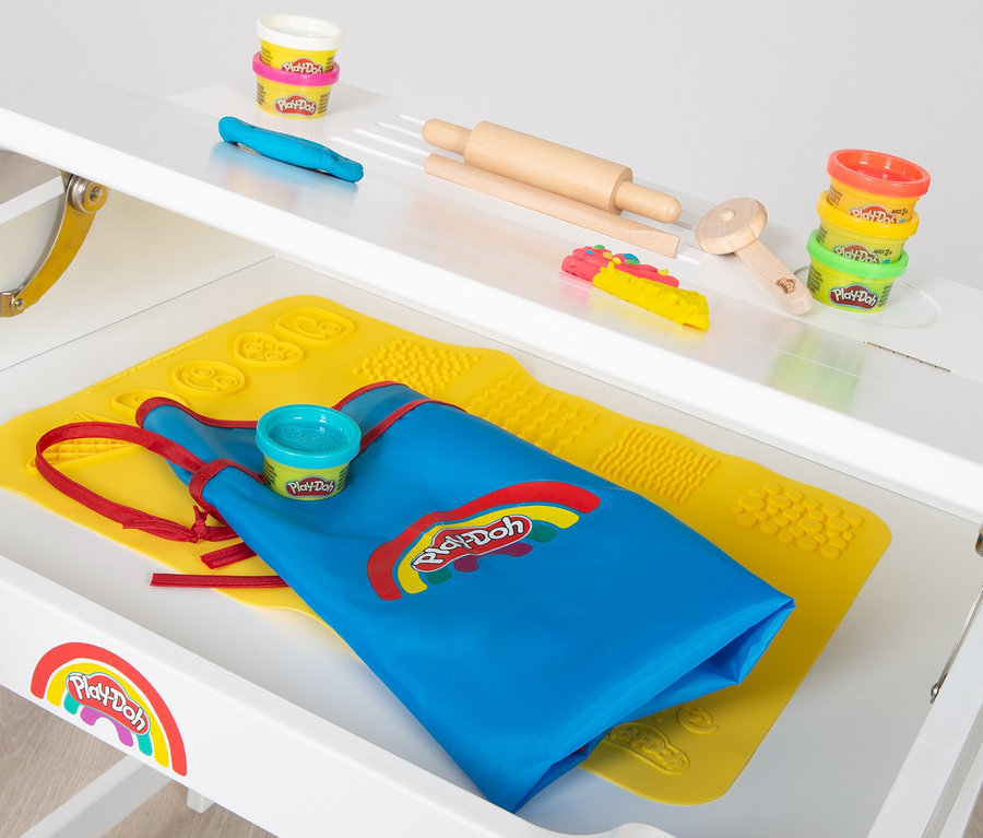 Auf einem weißen Tisch liegen Play-Doh-Dosen, Werkzeuge und eine blaue Schürze auf einer gelben Matte.