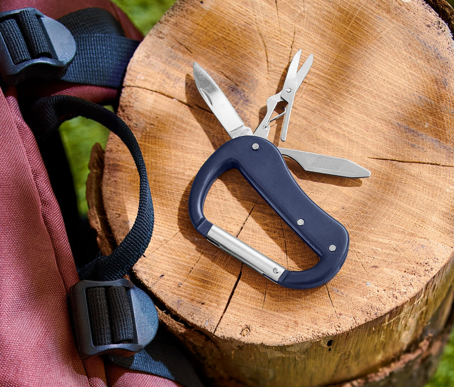 Ein blaues Multifunktionswerkzeug mit Karabiner liegt auf einem Baumstamm, daneben ein roter Rucksack.