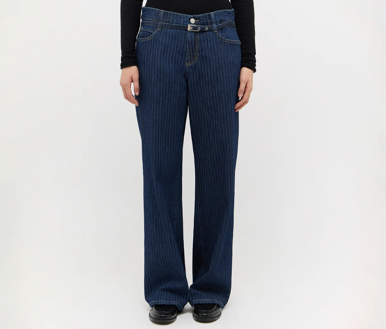 Eine Person trägt eine dunkelblaue, weit geschnittene Jeans mit dezenten vertikalen Streifen, ein schwarzes Langarmshirt und schwarze Schuhe. Die Jeans hat einen Gürtel mit zwei silbernen Ringen.