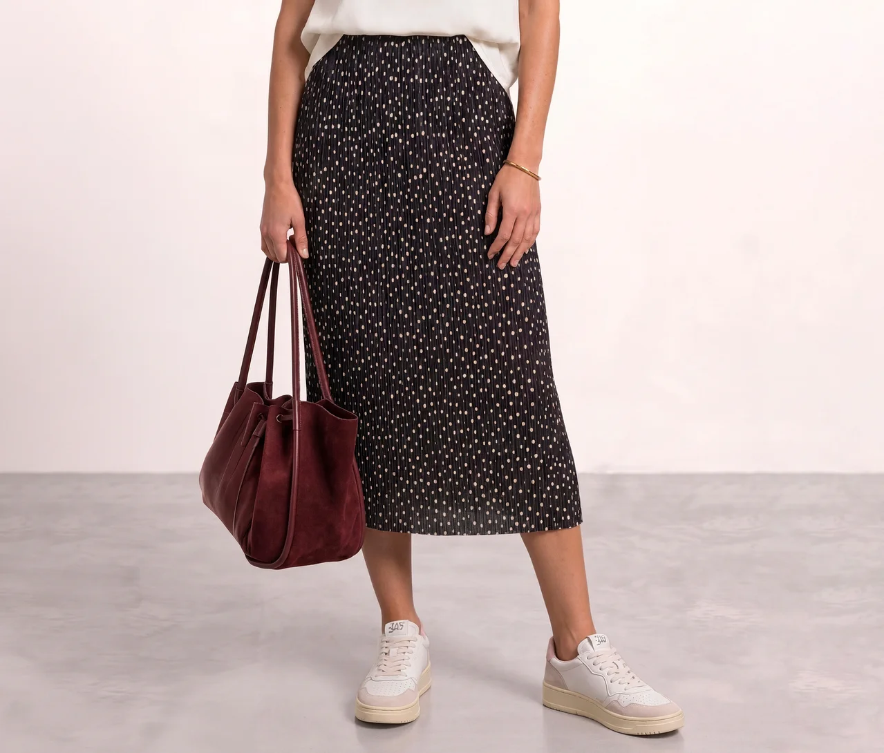 Eine Person trägt einen schwarzen, plissierten Street One Midi-Rock mit hellen Punkten, ein weißes Oberteil, weiße Sneaker und hält eine bordeauxrote Handtasche.