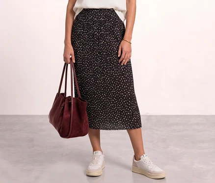 Eine Person trägt einen schwarzen, plissierten Street One Midi-Rock mit hellen Punkten, ein weißes Oberteil, weiße Sneaker und hält eine bordeauxrote Handtasche.