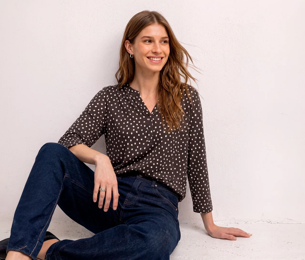 Frau sitzt lächelnd an einer weißen Wand, trägt ein dunkelbraunes Street One Blusenshirt mit weißen Punkten und dunkelblaue Jeans.