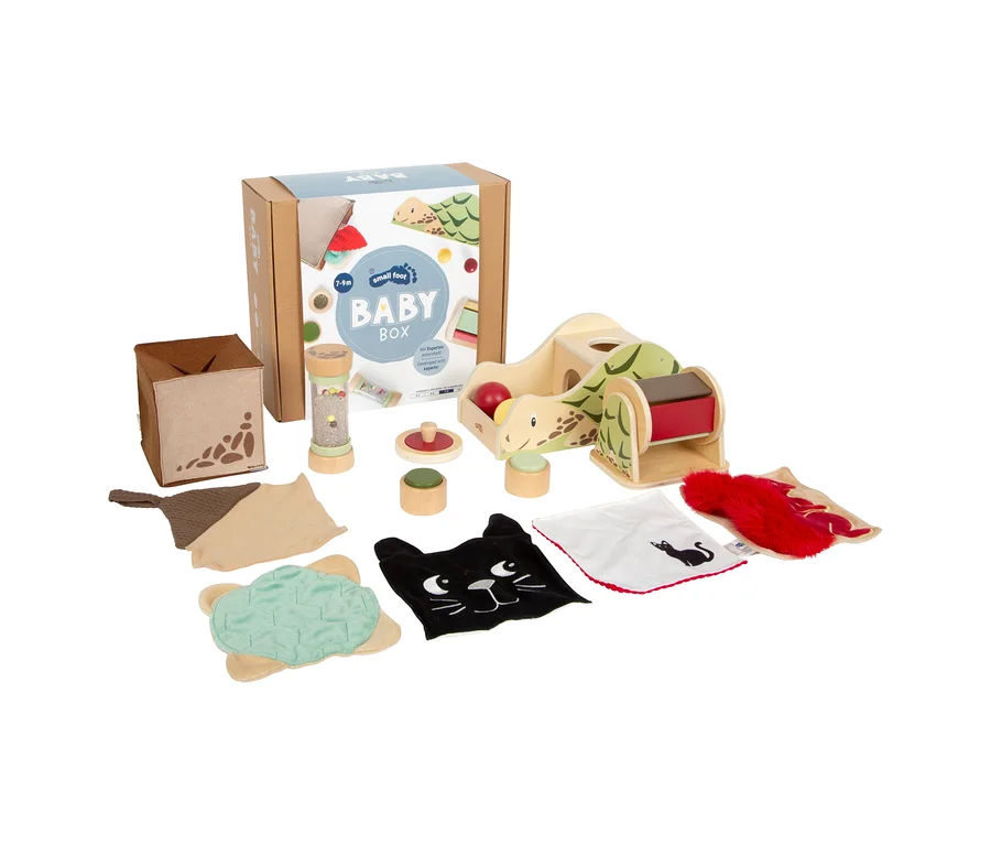small foot Baby-Box, 7–9 Monate, eine Kartonschachtel, umgeben von ihrem Inhalt: einem braunen Stoffwürfel, einer durchsichtigen Rassel, drei hölzernen Stapelspielzeugen, einem hölzernen Schildkrötenspielzeug mit rotem Ball und Rolle sowie mehreren Stoff-Sensorikpads, darunter eine grüne gesteppte Schildkröte, ein schwarzes Katzenkopf-Pad, ein weißes Katzensilhouetten-Pad und ein rotes flauschiges Pad.