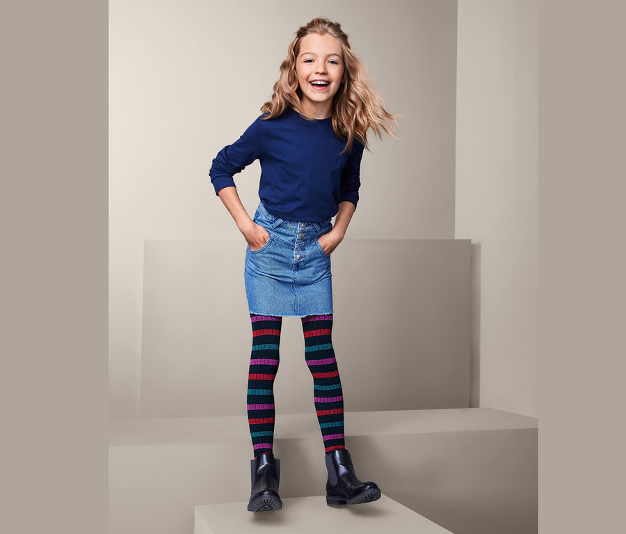 Lächelndes Mädchen posiert auf einem Podest mit Jeansrock, blauem Oberteil und 2 Strickleggings.
