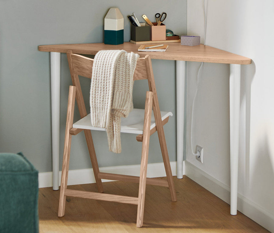 Ein Klappstuhl aus FSC®-zertifiziertem Eschenholz steht an einem dreieckigen Tisch. An der Wand hängt eine weiße Wandleuchte aus Metall.