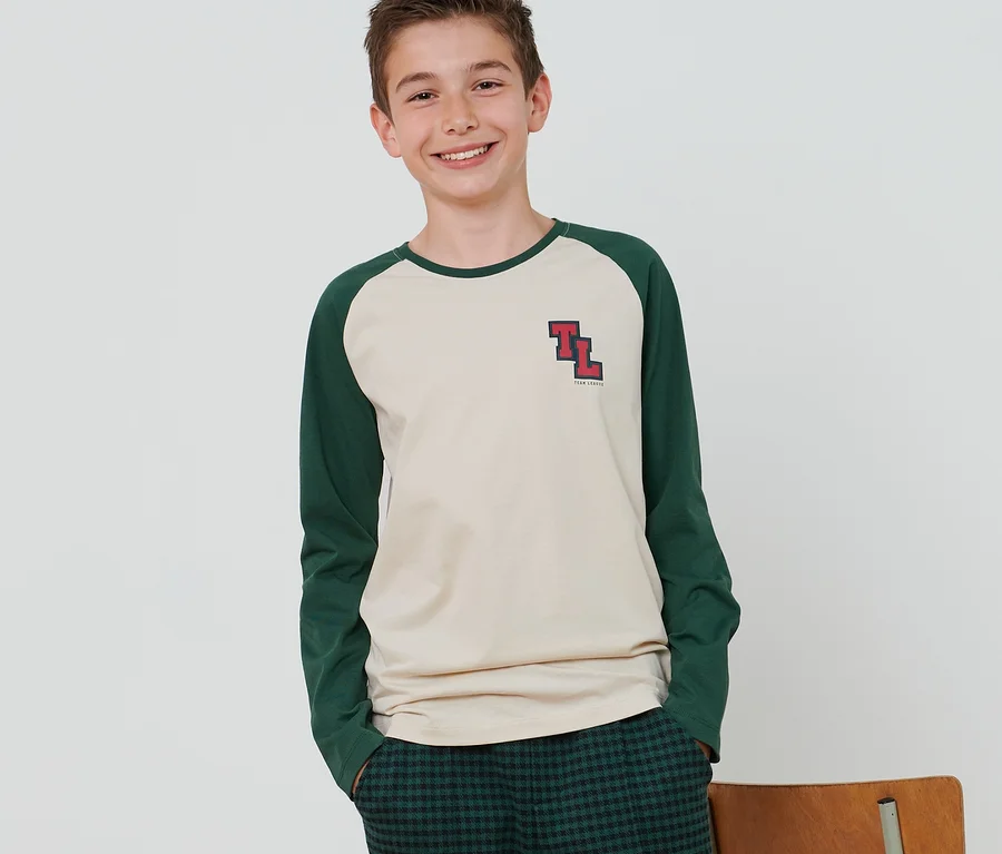 Ein Junge steht und trägt ein beiges Langarmshirt mit grünen Ärmeln, eine grüne Karohose und weiße Turnschuhe. Neben ihm steht ein Stuhl.