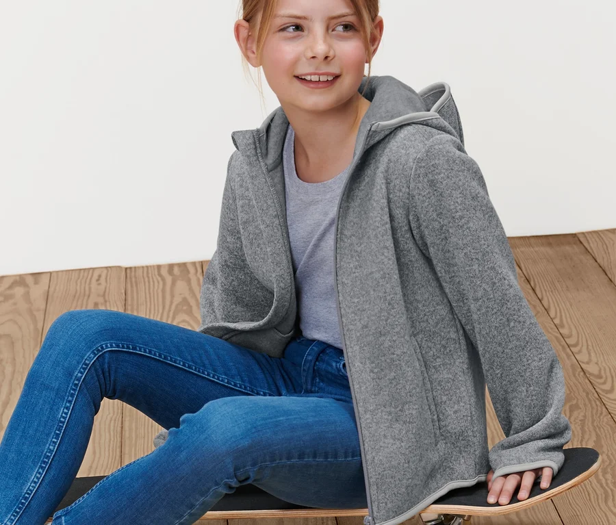 Mädchen sitzt auf einem Skateboard und trägt eine graue Kinder-Strickfleecejacke.