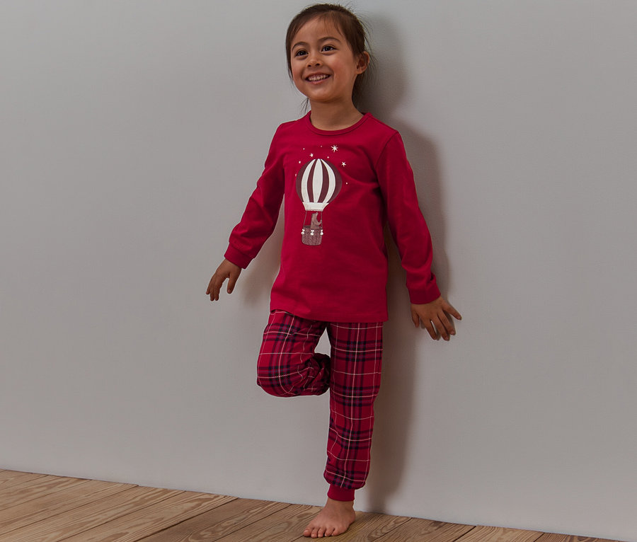 Lächelndes Mädchen steht an einer Wand, trägt einen roten Pyjama mit karierter Hose und einem Ballon-Aufdruck, der im Dunkeln leuchtet.