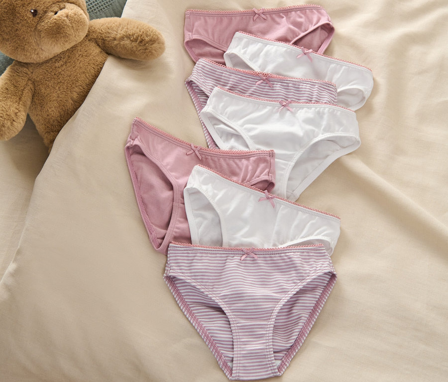 Sieben Kinder-Slips liegen neben einem Teddybär auf einem Bett.