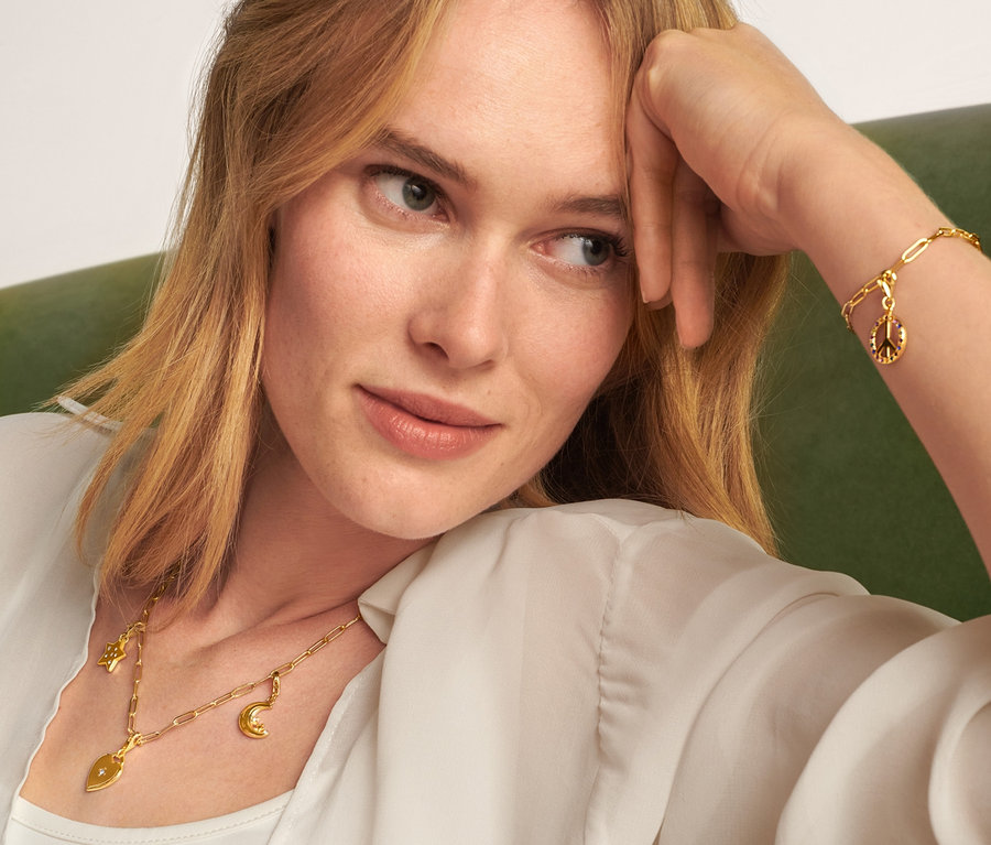 Blonde Frau sitzt auf einem grünen Sofa und trägt eine goldene Kette Collect, ein Armband Collect, ein Charm Set Love and Peace und ein Charm Set Moon and Stars.