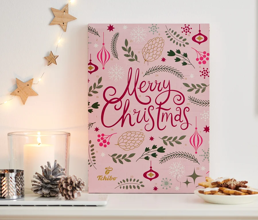 Tchibo-Adventskalender steht auf einer weißen Kommode neben einer Kerze, Deko und einem Teller mit Keksen.