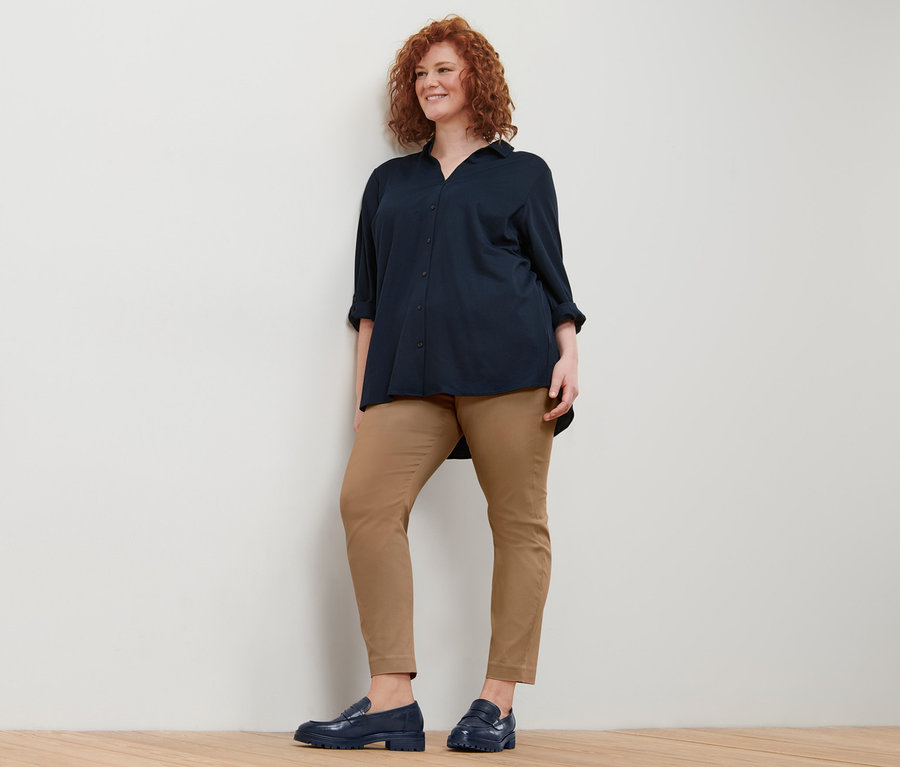 Frau mit lockigem, rotem Haar trägt eine dunkelblaue Piqué-Jerseybluse und eine Stretchhose.