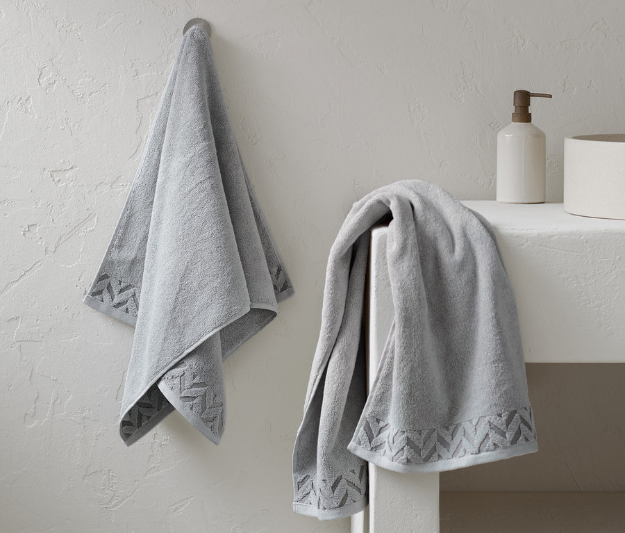 Zwei graue Premium-Handtücher liegen im Badezimmer. Eines hängt an der Wand und das andere liegt auf einem Regal.