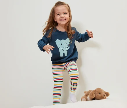Lächelndes kleines Mädchen steht auf einem Bett und trägt eine der 3 Kinder-Thermoleggings mit bunten Streifen.