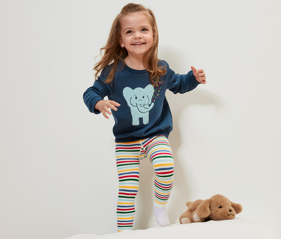 Lächelndes kleines Mädchen steht auf einem Bett und trägt eine der 3 Kinder-Thermoleggings mit bunten Streifen.
