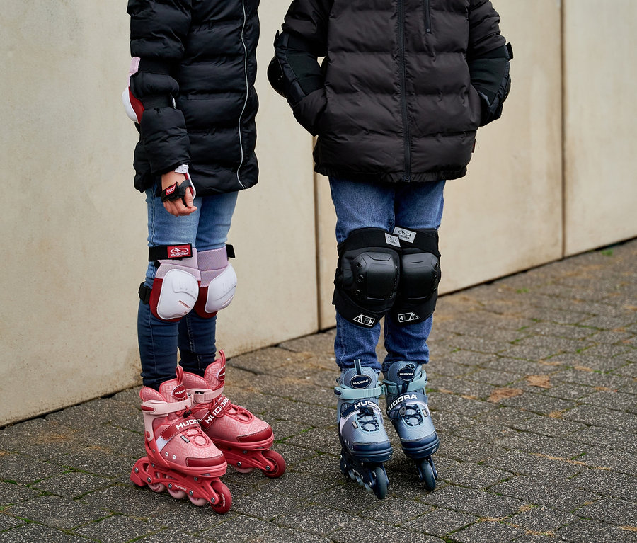 Zwei Kinder stehen draußen auf dem Bürgersteig und tragen Jacken, Jeans, Helme und Schoner sowie HUDORA Mia 2.0 Inlineskates.