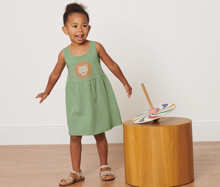 Mädchen trägt ein grünes Jerseykleid mit einem Löwen. Auf einem Hocker liegt ein Spielzeug.