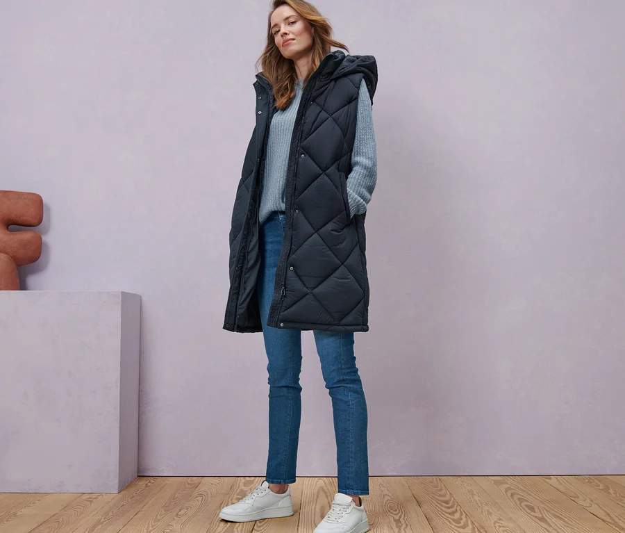 Frau steht in langer Steppweste mit Kapuze, rauchblauem Strickpullover und Skinny-Denim »Fit Hanna«.