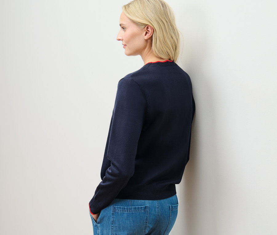 Blonde Frau steht seitlich, trägt einen blauen Feinstrick-Cardigan mit Wellenkante und Jeans.