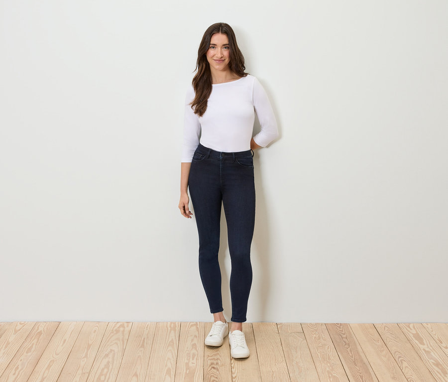 Eine Frau steht vor einer weißen Wand und trägt dunkelblaue Jeans, ein weißes Dreiviertelarmshirt und weiße Turnschuhe.