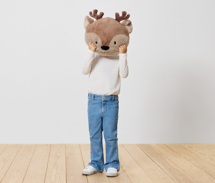 Ein Kind hält ein Stofftier-Rentier vor sein Gesicht, steht auf einem Holzboden vor einer weißen Wand.