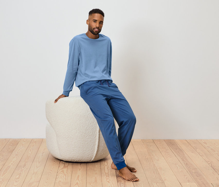 Mann sitzt auf einem weißen Pouf und trägt ein blaues Langarmshirt und eine blaue Jogginghose.