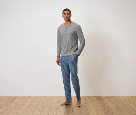 Mann steht auf einem Holzboden und trägt ein graues Langarmshirt und eine blau gestreifte Hose.