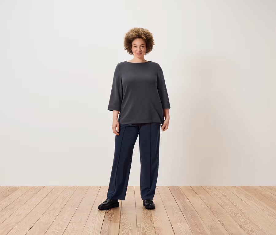 Eine Frau mit Afro-Frisur steht auf einem Holzboden und trägt eine graue Bluse und dunkelblaue Hosen.