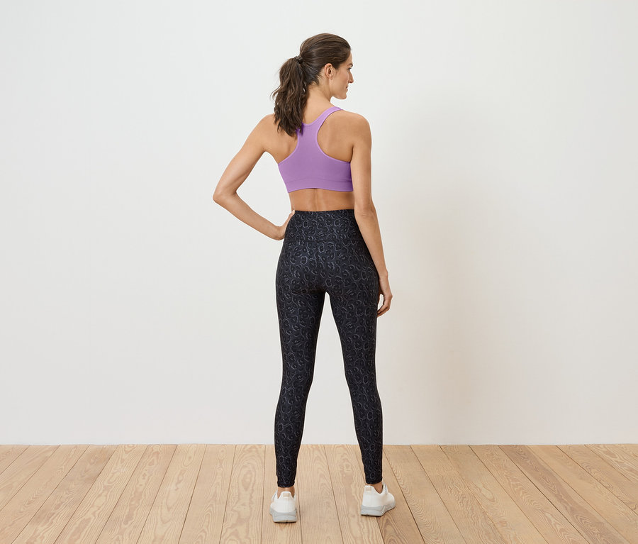 Frau steht mit dem Rücken zum Betrachter und trägt einen lila Sport-BH und schwarze Leggings.