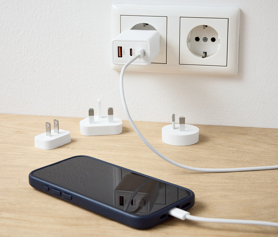 Ein weißer USB-Reiseadapter steckt in einer Steckdose und lädt ein Mobiltelefon auf. Auf dem Tisch liegen weitere Adapter.